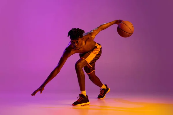 Молодой африканский мужчина, баскетболист в действии, движение с мячом изолированы на фиолетовом неоновом фоне. — стоковое фото