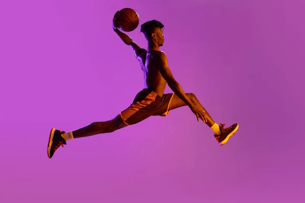 Fiatal afrikai férfi, kosaras akcióban, mozgó labda elszigetelt lila neon háttér. Oldalnézet — Stock Fotó