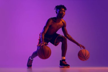 Genç Afrikalı bir adam, hareket halindeki basketbol oyuncusu iki topu mor neon arka planda izole edilmiş. Ön görünüm