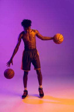 Genç Afrikalı bir adam, basketbol oyuncusu eğitimi almış, iki topu da mor neon arka planda. Tam boy portre