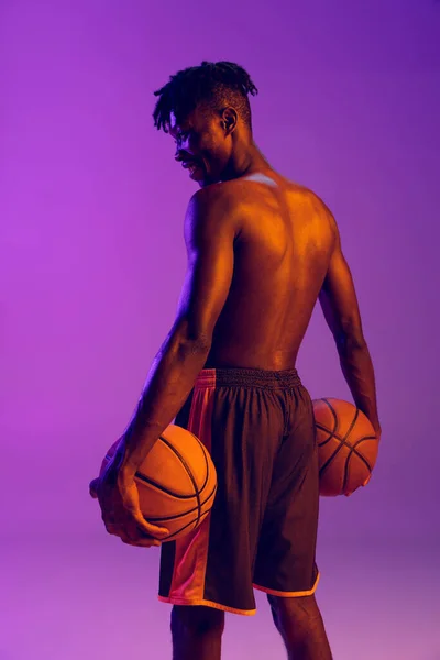 Homem africano jovem, jogador de basquete posando com duas bolas isoladas sobre fundo de néon roxo. Visão traseira — Fotografia de Stock