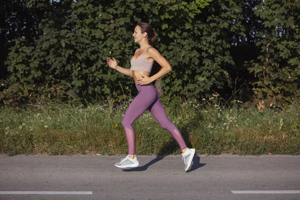 一个年轻而纤细的运动健美的高加索女人跑着，在宽阔的路上健身。侧视图 — 图库照片