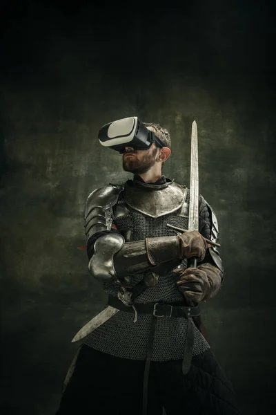 Portrait of one brutal bearded man, medeival warrior or knight in VR glasses with sword over dark background. — Stock Photo, Image