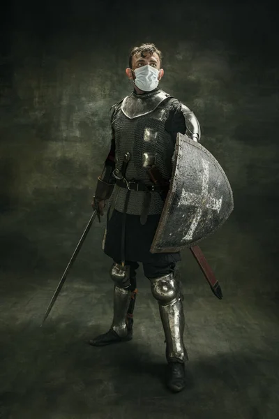 Ein brutaler bärtiger Mann, Medaillenkrieger oder Ritter in medizinischer Maske mit Schild und Schwert vor dunklem Hintergrund. — Stockfoto
