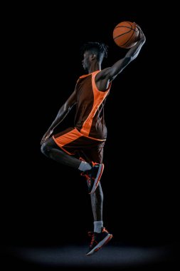 Dikey görüntü. Genç Afrikalı bir adam, basketbol oyuncusu topuyla atlıyor. Karanlık stüdyo geçmişinden izole edilmiş..
