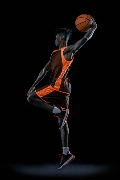 Immagine verticale. Giovane uomo africano, giocatore di basket che salta con palla isolata su sfondo scuro dello studio. — Foto Stock