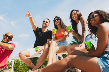 Bir grup genç, erkek ve kadın plajda dinleniyor, soğuk içecekler içiyor, eğleniyor, güneşleniyor.