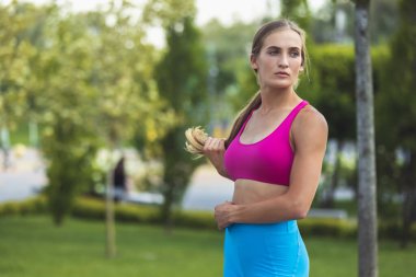 Parlak spor kıyafetleri içinde genç, zayıf, sportif beyaz bir kadın parkta ya da şehir caddesinde duruyor..