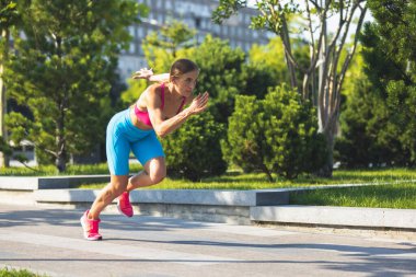 Yan görüş. Genç, zayıf, sportif beyaz bir kadın parkta ya da şehir caddesinde koşuyor..