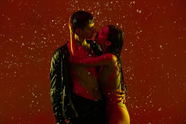 Casal jovem, homem e mulher abraçando, beijando sob a chuva sobre o fundo vermelho em luz de néon. — Fotografia de Stock