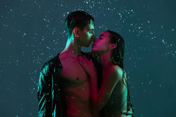 Casal jovem, homem e mulher abraçando, beijando sob a chuva sobre fundo azul em luz de néon. — Fotografia de Stock