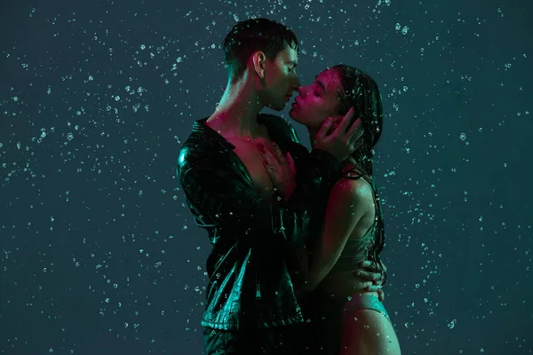 Casal jovem, homem e mulher abraçando, beijando sob a chuva sobre fundo escuro em luz de néon. — Fotografia de Stock