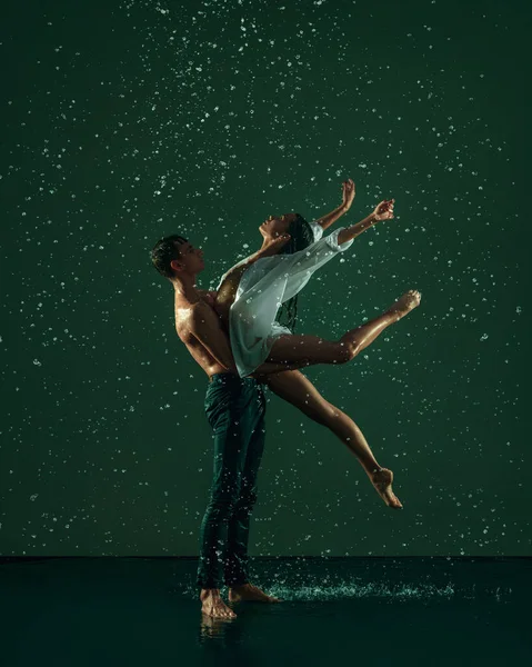 Two young ballet dancers, man and woman dancing in the raindrops over dark background. Art, action, inspiration concept.
