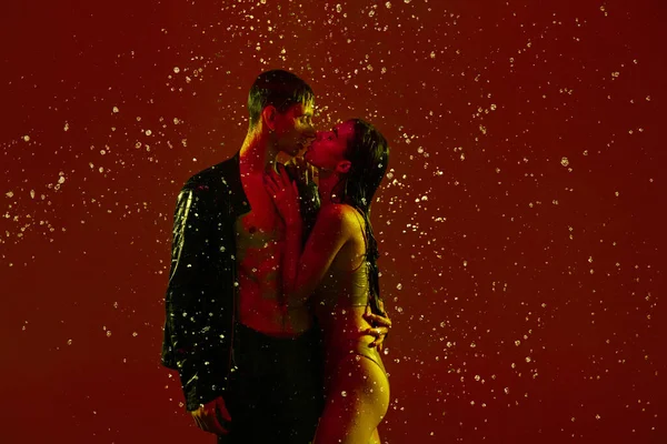 Młoda para, mężczyzna i kobieta przytulają się, całują się w deszczu na czerwonym tle w neonowym świetle. — Zdjęcie stockowe