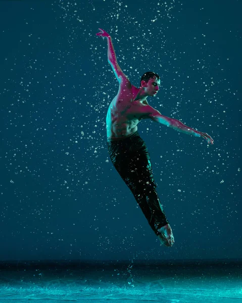 Een jonge mannelijke balletdanser in beweging, actie onder de regen over blauwe achtergrond in neon licht. — Stockfoto