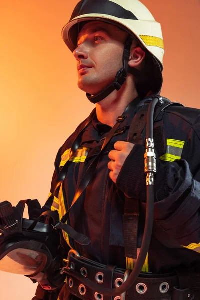 Ένας άνδρας πυροσβέστης ντυμένος με στολή ποζάρει πάνω από πορτοκαλί φόντο σε φώτα νέον. — Φωτογραφία Αρχείου