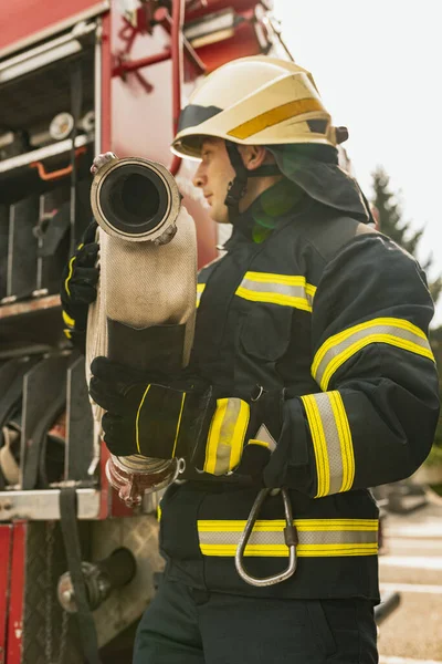 En ung manlig brandman klädd i uniform med vattenslang framför brandbilen. Närbild — Stockfoto