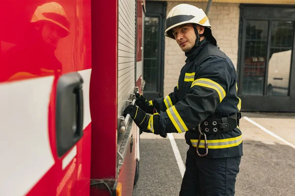 En ung manlig brandman klädd i uniform med skyddshjälm nära brandbil. — Stockfoto