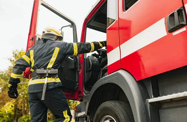 Ein junger Feuerwehrmann in Uniform mit Schutzhelm klettert in Feuerwehrauto. — Stockfoto