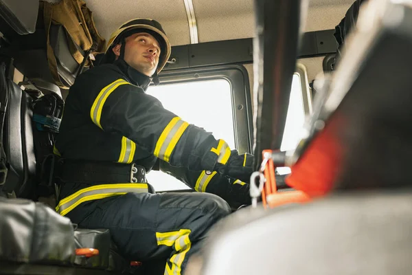 Un joven bombero vestido de uniforme con casco protector en camión de bomberos. —  Fotos de Stock