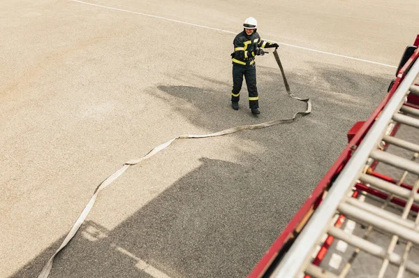 En ung manlig brandman klädd i uniform med vattenslang redo för utplacering — Stockfoto