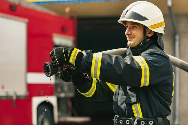 En ung manlig brandman i tjänst klädd i uniform med vattenslang släckning brand — Stockfoto