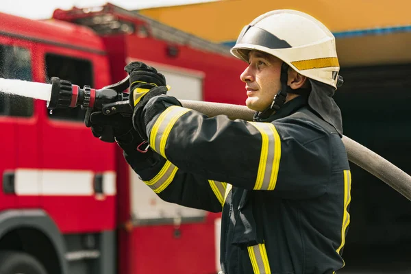 En ung manlig brandman i tjänst klädd i uniform med vattenslang släckning brand — Stockfoto