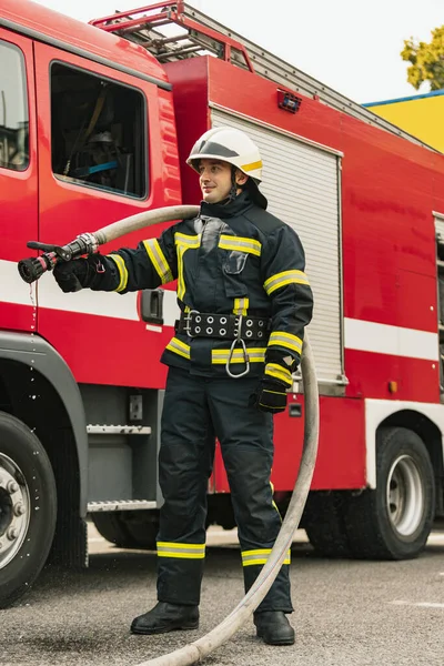 Ein junger Feuerwehrmann in Uniform mit Wasserschlauch am einsatzbereiten Feuerwehrauto — Stockfoto