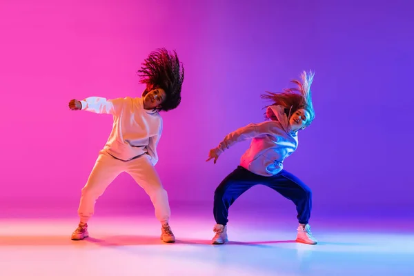 Two beautiful active girls dancing on gradient pink purple neon studio background — Stock Photo, Image