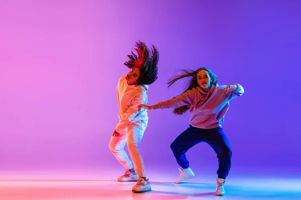 Deux belles jeunes filles dansant hip-hop dans des vêtements élégants sur fond dégradé coloré au néon. — Photo