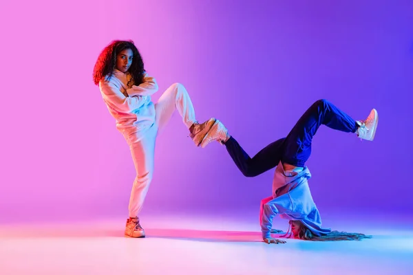 Dos hermosas chicas jóvenes bailando hip-hop dinámicamente sobre fondo degradado colorido en neón. — Foto de Stock