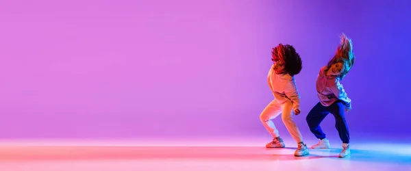 Twee mooie jonge meisjes dansen hip-hop dynamisch op kleurrijke gradiënt achtergrond in neon. — Stockfoto