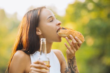 Yakından kesilmiş, elinde bira şişesi olan ve sıcak yaz pikniğinde sosisli yiyen genç kızın portresi.