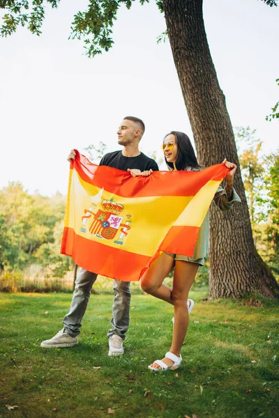 Повний портрет двох молодих футбольних уболівальників, чоловіка і жінки, які тримають прапор Іспанії, ізольований на зворотному боці парку. — стокове фото