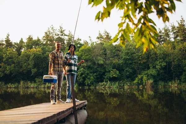Портрет молодой улыбающейся пары, держащей удочку и рыбалку в теплый летний день в сельской местности на закате — стоковое фото