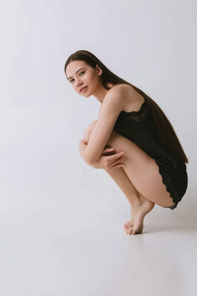 Full-lengths portrait of beautiful girl in black silk pajama sitting on grey studio wallpaper — Stock Photo, Image