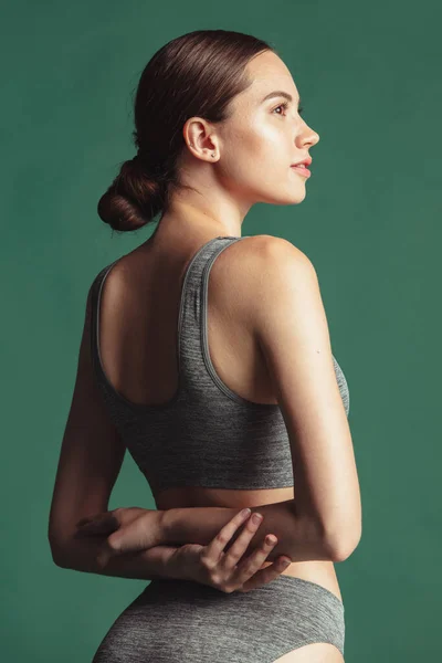 Retrato recortado. Joven hermosa mujer caucásica en ropa interior gris mirando hacia otro lado aislado sobre fondo de estudio verde. Vista trasera — Foto de Stock