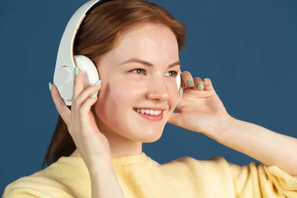 Retrato de cerca de una joven pelirroja con auriculares mirando hacia otro lado aislada sobre fondo azul. — Foto de Stock