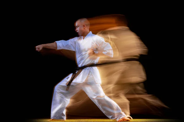 Full-length portrait of young karate sportsman wearing white kimono isolated over black background. Mixed light effect — Stock Photo, Image