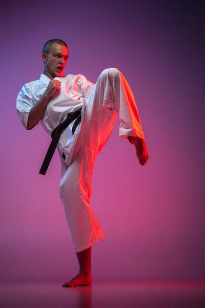 Fullständigt porträtt av kaukasisk karate, judo, taekwondo idrottsman i vit kimono träning isolerad på lutning rosa lila bakgrund i neonljus — Stockfoto