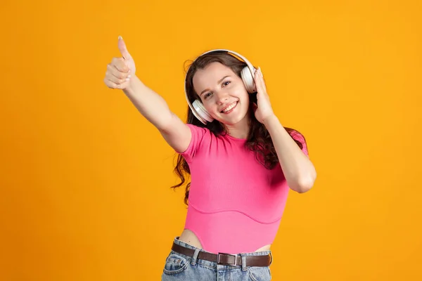 Portrait recadré de jeune belle fille écoutant de la musique dans les écouteurs avec les pouces vers le haut isolé sur fond de studio orange — Photo