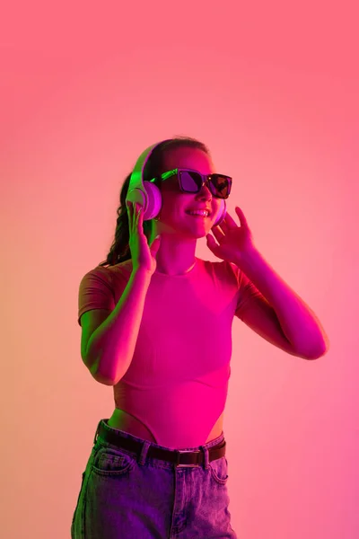 Immagine verticale ritagliata di giovane ragazza sorridente in cuffie e occhiali da sole isolato su sfondo studio gradiente in luci al neon — Foto Stock