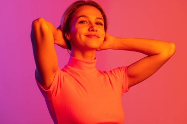 Genç ve güzel bir kız kameraya hafifçe gülümsüyor ve neon ışıklarıyla izole edilmiş saçlarını düzeltiyor.