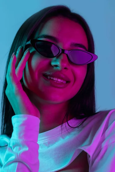 Ritratto ravvicinato di giovane bella ragazza in posa con occhiali da sole isolati su sfondo blu in luce al neon. Bellezza — Foto Stock