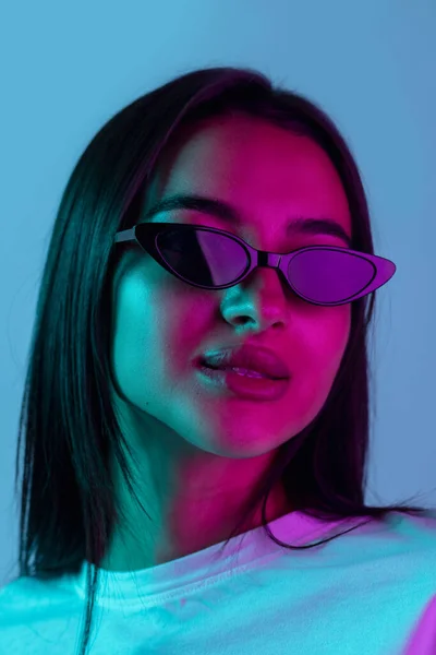 Ritratto ravvicinato di giovane bella ragazza in posa con occhiali da sole isolati su sfondo blu in luce al neon. Trendy — Foto Stock