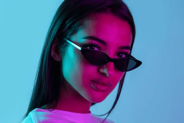 Close-up ritratto di giovane bella ragazza in posa in occhiali da sole isolati su sfondo blu in luce al neon. — Foto Stock