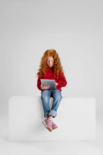 Teljes hosszúságú portré a kis göndör mosolygós lány, gyermek ül és gépel tabletta elszigetelt szürke stúdió háttér. — Stock Fotó