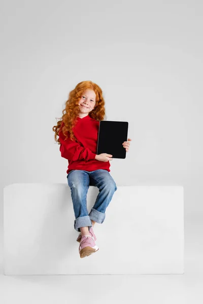 Portrait of little red-haired curly girl, child pointing at tablet isolated over gray studio background. — Stock Photo, Image