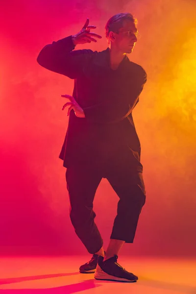 Młody biały mężczyzna w stylowe czarne ubrania taniec hip-hop izolowane nad gradient różowy żółty tło w neon światło w dym — Zdjęcie stockowe