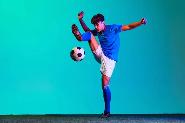 Retrato horizontal de larga duración del joven deportista, futbolista en movimiento pateando una pelota aislada sobre fondo azul — Foto de Stock